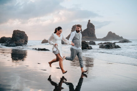 Los novios corren descalzos de la mano por la orilla, reflejados en la arena mojada, ante unos roques