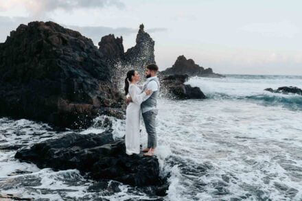 Los novios se abrazan sobre una roca negra mientras una ola rompe detrás, entre roques y espuma