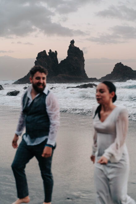 Los novios, con la ropa mojada, caminan riendo por la arena, desenfocados ante unos roques en el mar