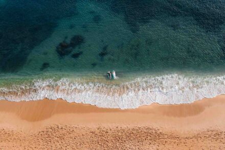 Los novios tumbados en el agua turquesa, justo detrás de la espuma de las olas que llegan a la arena dorada, desde el aire