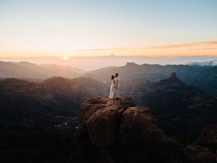 Los novios de pie sobre una gran roca en lo alto de las cumbres, con valles a sus pies y el sol en el horizonte