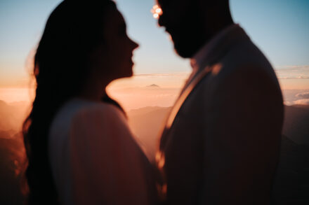 Los novios, en silueta desenfocada, frente a frente sobre un horizonte de montañas y nubes a contraluz
