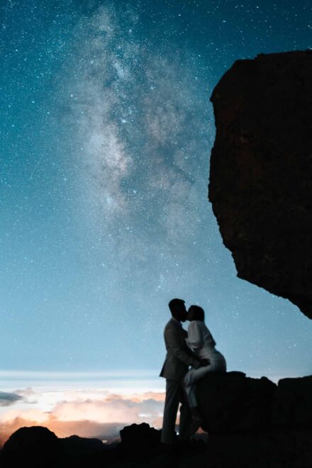 Los novios se besan en silueta bajo la Vía Láctea, ella sentada en una roca, sobre un mar de nubes
