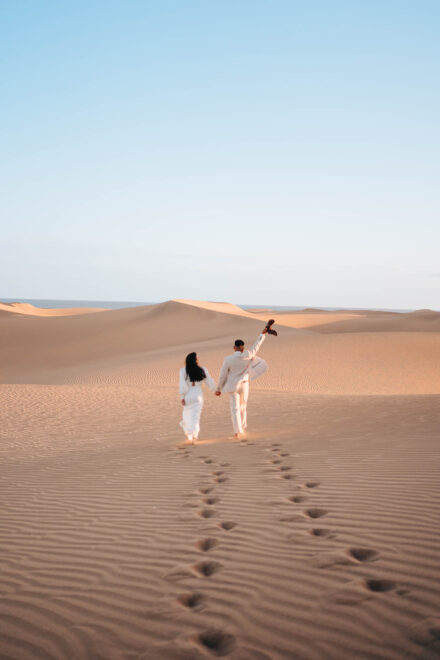 Los novios, vistos de espaldas, caminan de la mano por las dunas, él con los zapatos en alto, dejando sus huellas