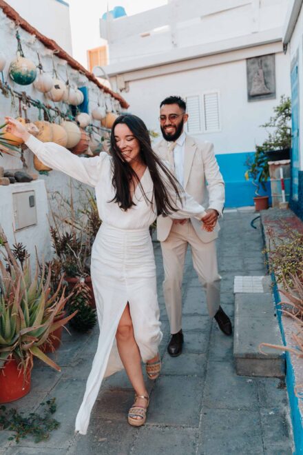 La novia ríe con el pelo al viento y tira del novio por un callejón blanco con boyas colgadas en la pared