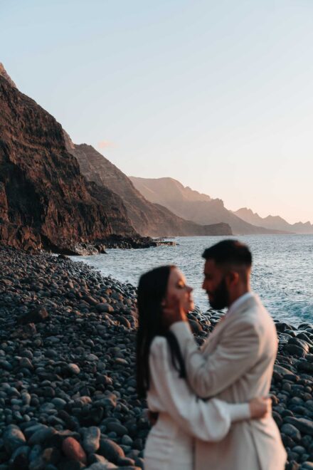 Los novios, desenfocados, se abrazan en primer plano ante los acantilados y el mar bajo un cielo rosado