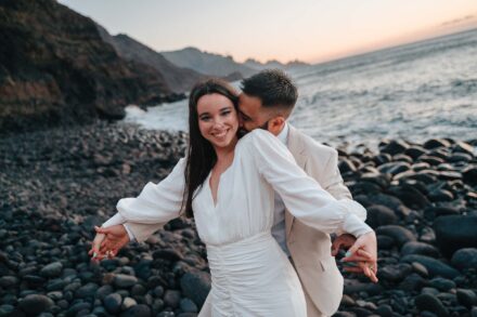 El novio abraza por detrás a la novia, que sonríe con los brazos abiertos en una playa de cantos rodados