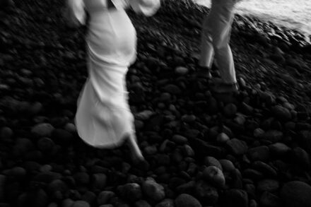 Los novios caminan movidos por una playa de cantos rodados, vistos de cintura para abajo, en blanco y negro