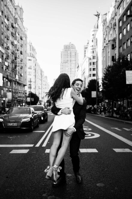 El novio, riendo, levanta en brazos a la novia en mitad de la Gran Vía de Madrid, en blanco y negro
