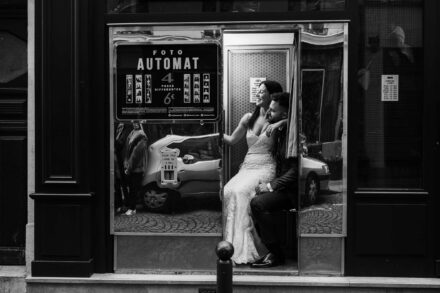 Los novios sentados dentro de una cabina de fotomatón, ella riendo con el brazo sobre él, en blanco y negro