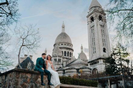 Los novios sentados en un muro de piedra, sonriendo bajo la basílica del Sacré-Cœur de París
