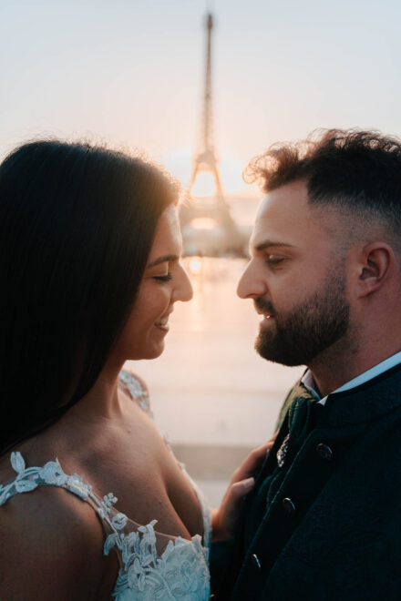 La novia sonríe con los ojos bajos frente al novio, con la torre Eiffel desenfocada al fondo