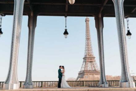 Los novios se besan entre las columnas de hierro de un puente, con la torre Eiffel al fondo, en París