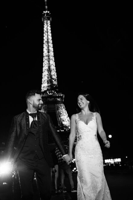 Los novios caminan de la mano riéndose de noche, con la torre Eiffel iluminada detrás