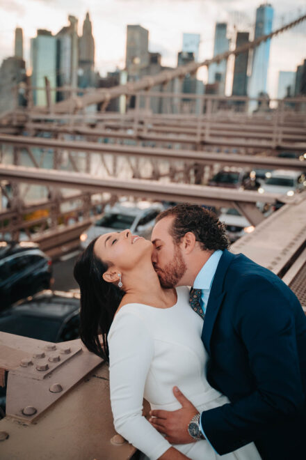 El novio besa el cuello de la novia, que ríe con los ojos cerrados sobre la pasarela de un puente de hierro