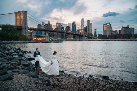 El novio sujeta la cola del vestido mientras la novia camina por las rocas junto al puente de Brooklyn