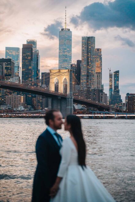 Los novios, desenfocados, a punto de besarse frente al puente de Brooklyn al anochecer