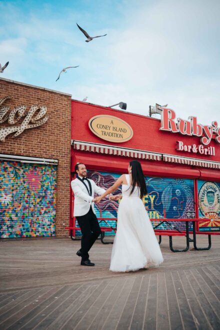 Los novios bailan de la mano en el paseo de madera junto al mar, bajo las gaviotas, ante un bar rojo con murales