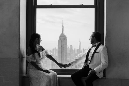 Los novios se dan la mano sentados en una ventana con el Empire State detrás, en blanco y negro