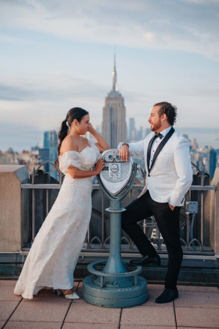 Los novios se miran en su postboda, apoyados en unos prismáticos de mirador con el Empire State al fondo