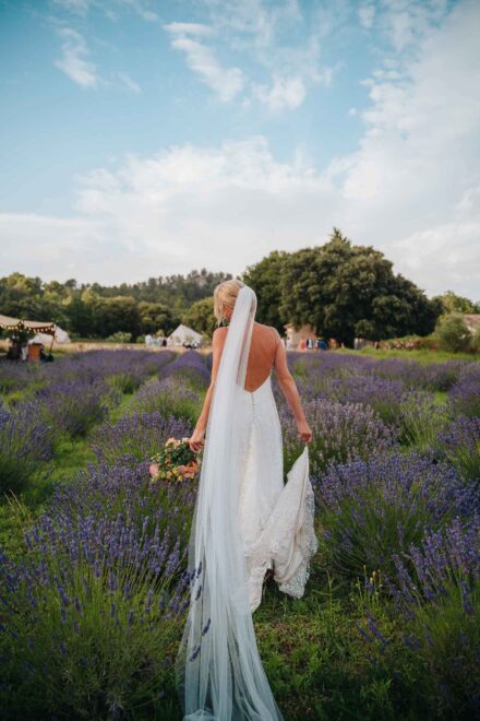 La novia, de espaldas y con un velo largo, camina por un campo de lavanda hacia unas carpas con invitados