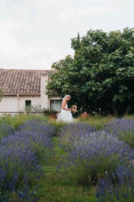 La novia mira su ramo entre hileras de lavanda, junto a una casa con tejado de teja y una higuera
