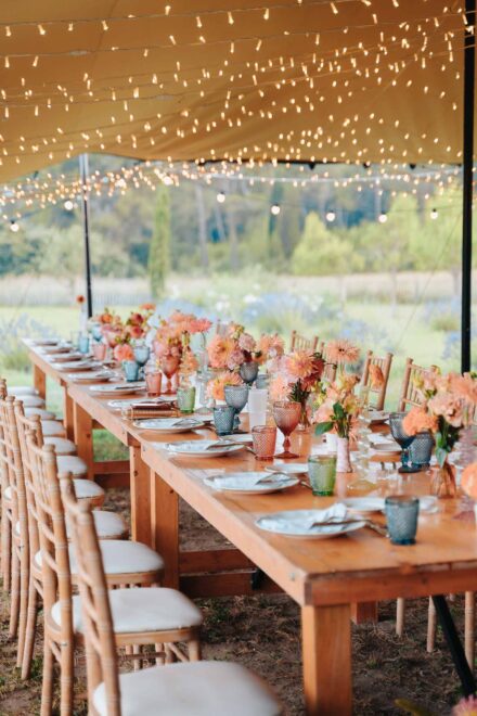 Mesa de madera bajo una carpa con luces, con dalias coral y vasos de colores, frente a un campo