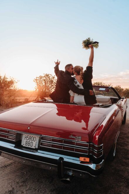 Los novios se besan en un descapotable rojo antiguo al atardecer; ella levanta el ramo al cielo