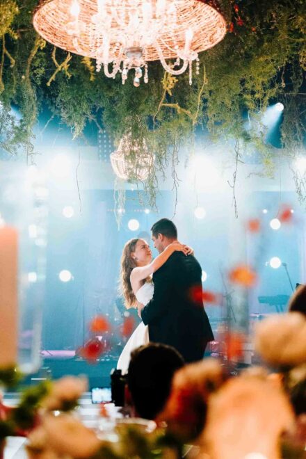 Los novios bailan abrazados bajo una lámpara de araña entre plantas colgantes, vistos tras los centros de flores