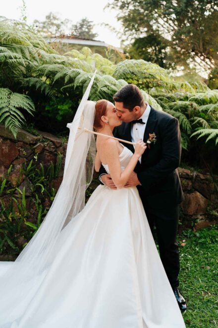 Los novios se besan frente a unos helechos; la novia sostiene una sombrilla blanca y el velo cae hasta el suelo