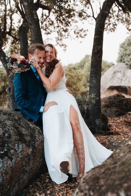 La novia, en las rodillas del novio sobre una roca, le coge la cara y los dos ríen con los ojos cerrados