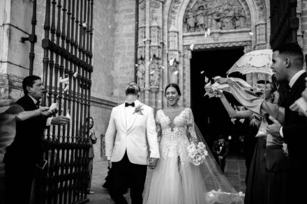 Los novios salen de la mano por una portada gótica entre una lluvia de pétalos, en blanco y negro