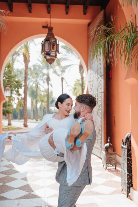 El novio lleva en brazos a la novia, con zapatos azules, bajo un arco de estilo árabe con un farol colgado