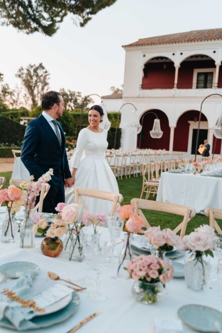 Los novios, de la mano, entre las mesas del banquete con jarrones de rosas, frente a una casa blanca de arcos rojos