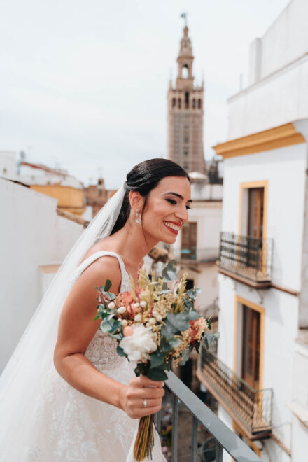 La novia sonríe con el ramo, asomada a un balcón, con la Giralda de Sevilla al fondo