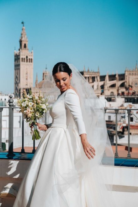 La novia, con vestido de manga larga y velo, posa con el ramo en una azotea frente a la Giralda y la catedral de Sevilla