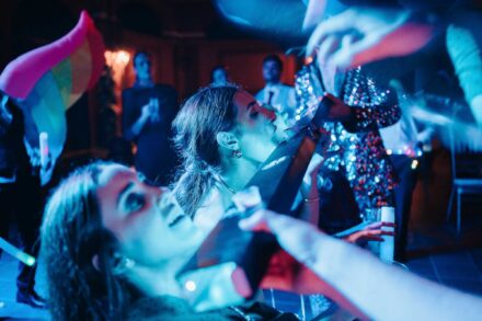 Dos invitadas echan la cabeza atrás para beber un chupito bajo luz azul, con una bandera arcoíris al lado