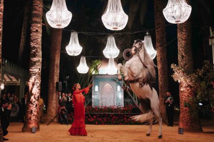 Una bailaora con vestido rojo alza el brazo ante un caballo blanco encabritado, bajo lámparas de araña entre palmeras