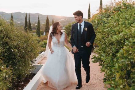 Los novios pasean de la mano y se sonríen por un camino junto a unos naranjos, con cipreses y montañas al fondo