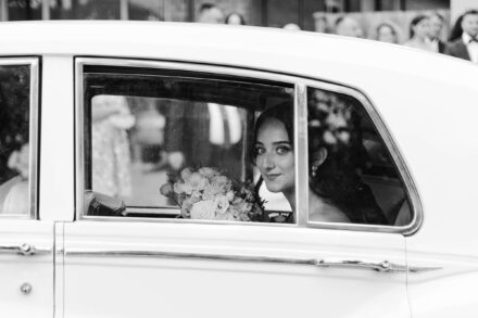 La novia mira a cámara tras la ventanilla de un coche antiguo blanco, con el ramo al lado, en blanco y negro