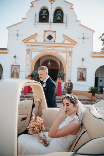 La novia se lleva una mano a los ojos, sonriendo, en un descapotable crema frente a una ermita con espadaña