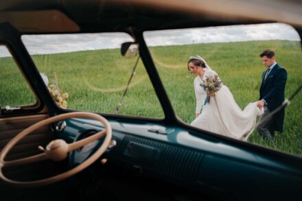 Los novios caminan por un campo de cereal verde, vistos a través del parabrisas de una furgoneta antigua