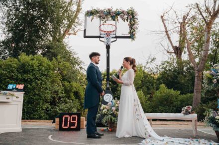La novia lee sus votos con un micrófono frente al novio, bajo una canasta de baloncesto adornada con flores