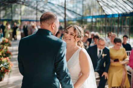 La novia sonríe al novio, de espaldas y con chaqueta verde, durante la ceremonia en el invernadero