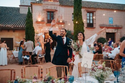 Los novios entran saludando al banquete en el patio de una hacienda, ella con el ramo, entre sillas de bambú