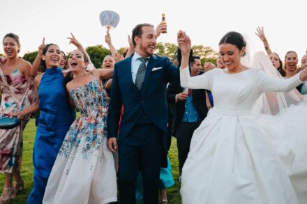Los novios bailan con las manos en alto en el césped, rodeados de invitadas que gritan y levantan los brazos