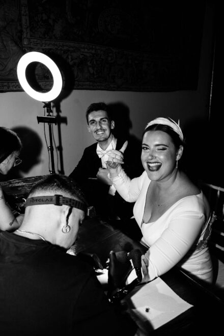 La novia guiña un ojo mientras le tatúan el antebrazo junto a un aro de luz, en blanco y negro