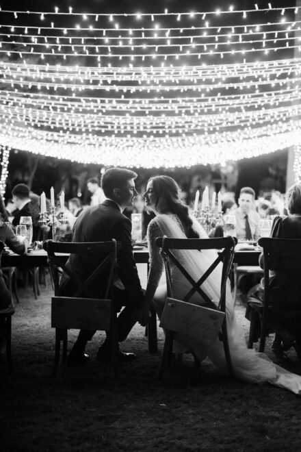 Los novios, de espaldas en la mesa del banquete, se miran bajo un techo de guirnaldas de luces, en blanco y negro