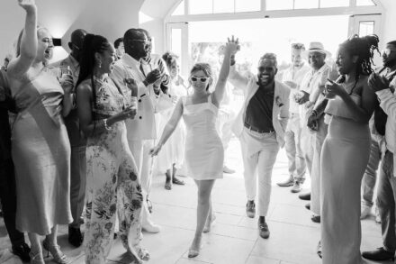 Los novios entran de la mano entre invitados que ríen y aplauden, ella con vestido corto y gafas blancas