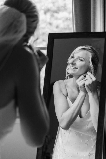 La novia sonríe reflejada en un espejo de pie mientras se pone los pendientes, en blanco y negro